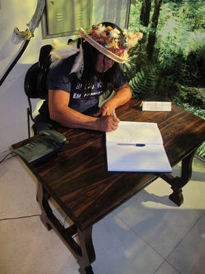 Indio usando calça comprida e blusa, portando coca, sentado, assinando livro de assintaura da exposição sobre uma mesa 